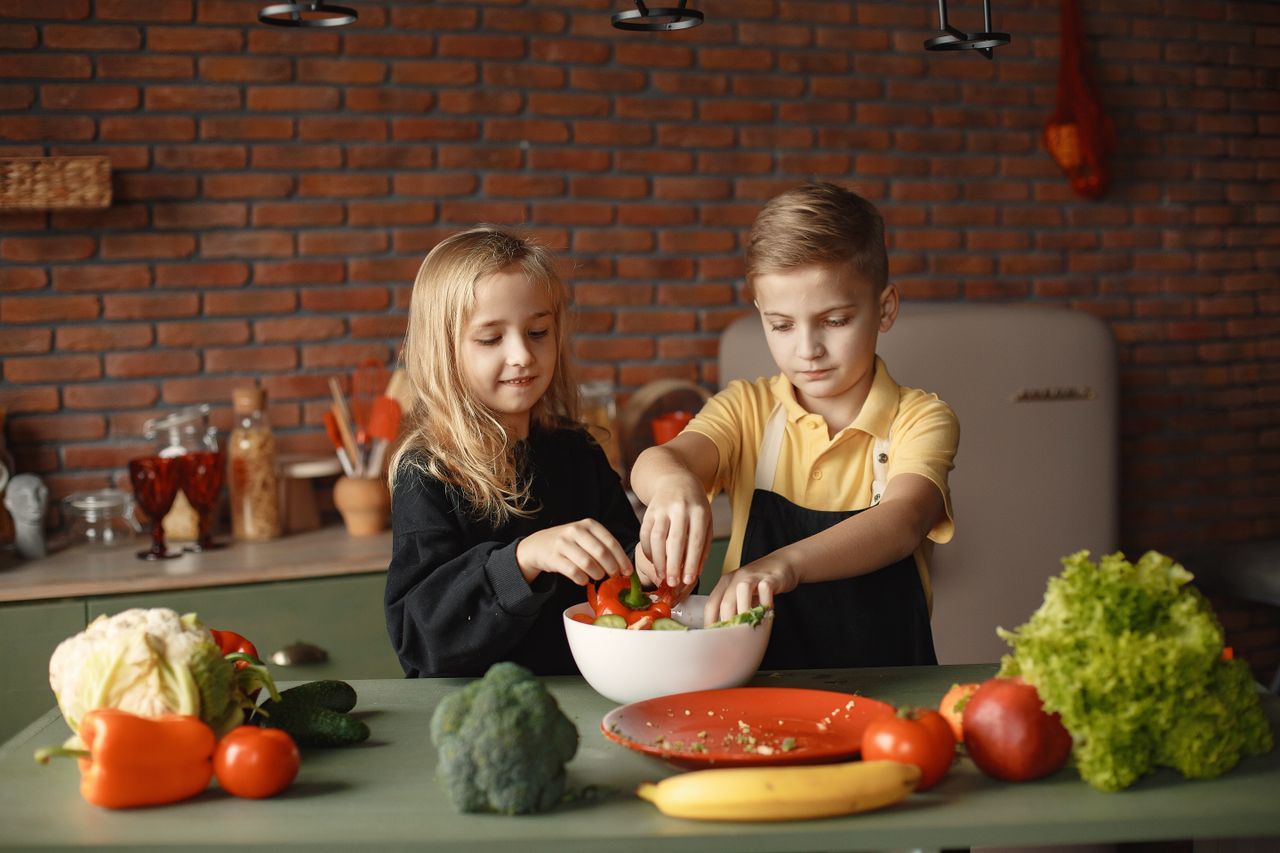 Cheerful girl helping little boy in apron preparing salad while standing in kitchen near table with mix of fresh vegetables behind brick wall