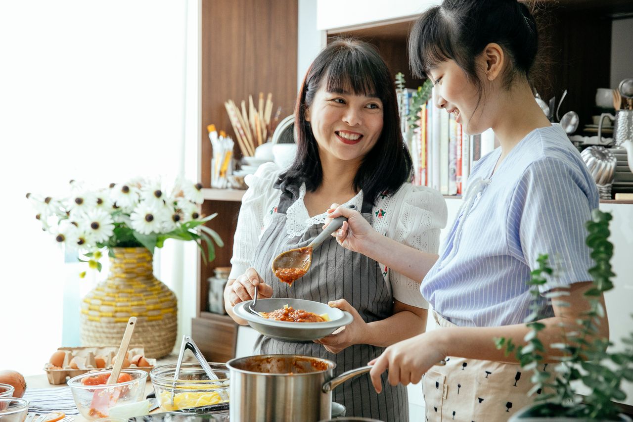 Cheerful Asian female housewives in aprons adding sauce in pasta while cooking together in kitchen