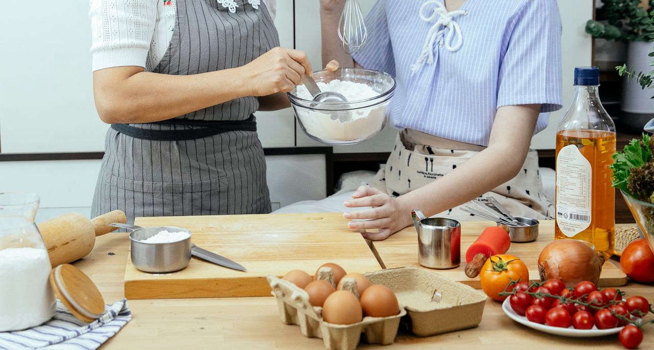 journey-01 Unrecognizable female cooks with bowl of flour standing at table with cutting board and various ingredients in kitchen while making dough