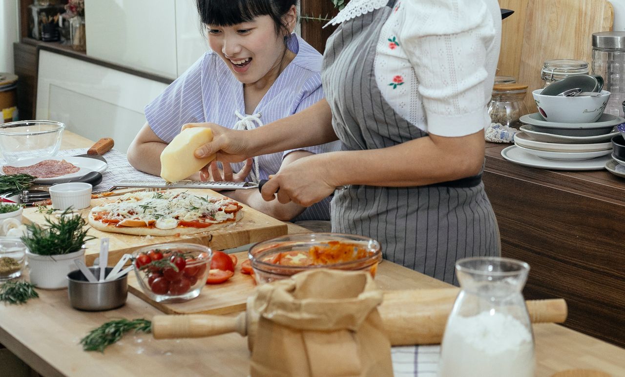 Happy Asian woman at table with unrecognizable woman rubbing cheese on uncooked pizza in kitchen with various ingredients during cooking process